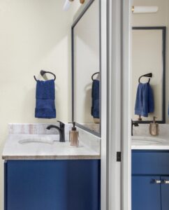 A modern bathroom features a blue vanity with a white marble countertop. A blue towel hangs on a black towel ring. A tan soap dispenser sits beside the black faucet. A large mirror reflects the scene, brightening the neutral-toned walls.
