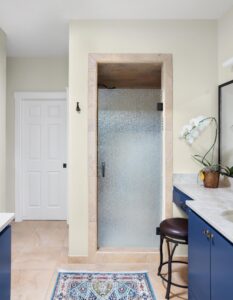 Modern bathroom with a frosted glass shower door framed by beige tiles. Blue vanity with a marble countertop, sink, and orchid plant. A stool sits nearby. White door in the background, and a patterned rug on the light tile floor.