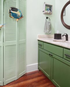 A bathroom with mint green walls and dark green cabinets. There's a round mirror above a countertop with a tissue box. A decorative fish hangs on a louvered door, and a towel is on a wall hook. Wood flooring completes the design.
