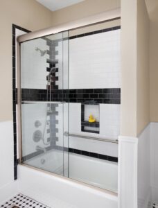 Modern bathroom with a glass-enclosed shower/bathtub combo. White subway tiles with black accent stripes line the walls. A small niche holds a yellow rubber duck. Black and white checkered tile floor enhances the classic look.