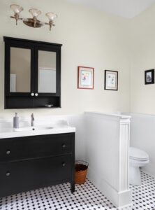 A bathroom with a black vanity and white sink, topped by a wall-mounted mirror cabinet. A toilet is enclosed by a partial wall. Black and white tiles cover the floor. Small framed artworks hang on the walls, and a wicker basket is on the floor.
